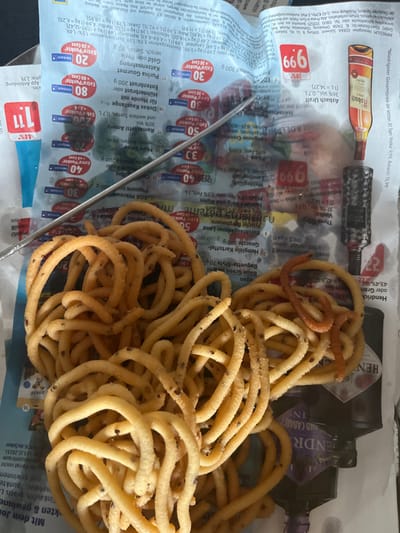 The image depicts an indoor scene in Tuttlingen, Germany, presenting a static arrangement of items on a surface covered by a German supermarket flyer.

**Scene Elements:**
*   **Base Layer:** The immediate surface is covered by an open multi-page advertisement flyer, featuring "EDEKA" branding and German text. Advertisements for "Purina Gourmet Katzenahrung" (cat food), "Hühnerbrust" (chicken breast), "Ramazzotti Amaro," "Asbach Uralt," "Vodka," and "Hendrick's Gin" are visible, along with pricing and "Payback" loyalty program details.
*   **Primary Subject (Food):** A large, irregularly shaped pile of light beige to golden-brown fried snacks, likely a variety of Murukku, is centrally positioned. These snacks are tubular and coiled, and contain small, dark, embedded seeds. One distinct piece of a reddish-orange fried snack is intermingled within the pile on the right side.
*   **Objects on Flyer:**
    *   A thin, silver-colored metallic skewer or probe lies diagonally across the upper-middle section of the flyer, partially resting on the snacks.
    *   Two dark-colored bottles are partially visible on the right side, underneath and beside the snacks. One bottle is brown with a label indicating "Asbach Uralt." The second, largely obscured, shows part of a label with text consistent with "Hendrick's Gin."
    *   Small, blurred green specks are visible in the background, near the top of the flyer, beneath the skewer.
*   **People:** No individuals are present or visible in the frame.
*   **Actions/Interactions:** All depicted items are stationary and arranged on the flyer.