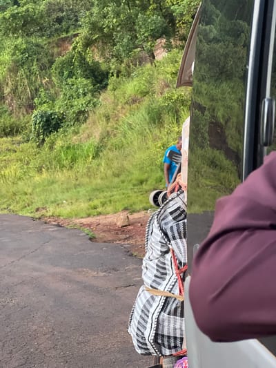 The image depicts a roadside scene with lush green vegetation and a dirt road in the foreground. The road is cracked and appears to be unpaved in some sections. On the side of the road, there are several large bags with a black and white checkered pattern, tied with red and tan ropes. In the background, a man wearing a blue shirt is visible, standing near the bags. He appears to be involved in some activity related to the bags or transportation. The reflection on a nearby vehicle's surface shows the surrounding green landscape. The overall impression is of a rural or semi-rural setting.