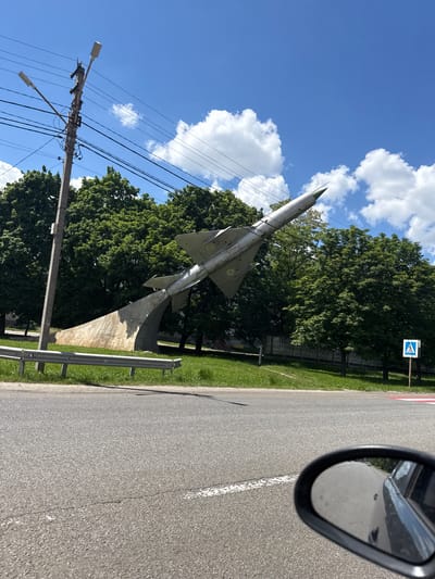 The image depicts a roadside scene in Chuhuiv, Ukraine, under bright daylight conditions. The central subject is a monument featuring a silver-grey fighter jet, likely a MiG, mounted on a slanted concrete pedestal, positioned to appear as if taking off. The jet bears distinct yellow and blue Ukrainian roundels on its tail fin.
The scene is viewed from inside a vehicle, with a black-framed side mirror visible in the upper right, reflecting a portion of the asphalt road and potentially the car itself. To the right of the jet, a paved road stretches, featuring a solid white line and a red and white striped curb. A metal guardrail runs alongside the road, separating it from a strip of green grass and a dense line of large, leafy green trees, indicative of summer or late spring.
Above the tree line, a bright blue sky is adorned with scattered white cumulus clouds. In the upper right background, a blue square sign with a white triangle and a pedestrian symbol (indicating a pedestrian crossing) is visible. On the far left, a utility pole with multiple power lines and two streetlights stands tall against the sky. The overall impression is a clear, sunny day with good visibility, showcasing a civic monument alongside a typical road.