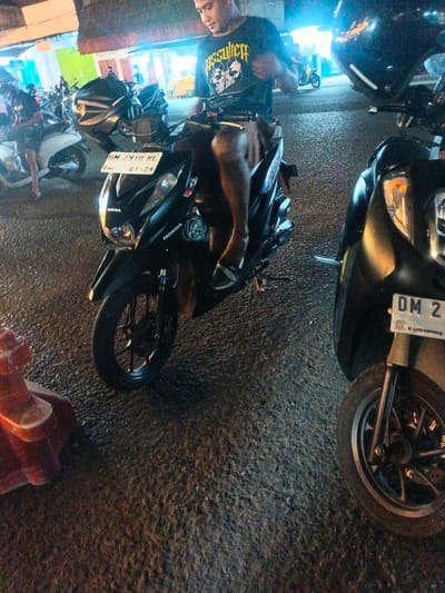 The image depicts a nighttime scene on a paved road in Luwoo, Indonesia, likely a commercial or pedestrian area based on the illuminated storefronts in the background. The foreground is dominated by two black scooters. A person, partially visible and wearing a black t-shirt with a graphic print, is seated on the scooter on the left. The scooter's license plate is visible and reads "DM 2410 RC". The scooter on the right also has a license plate, partially obscured but showing "DM 2". Other scooters and individuals are visible further in the background, suggesting a gathering or busy street. The lighting is dim, with artificial light sources illuminating the scene, creating reflections on the wet pavement.
