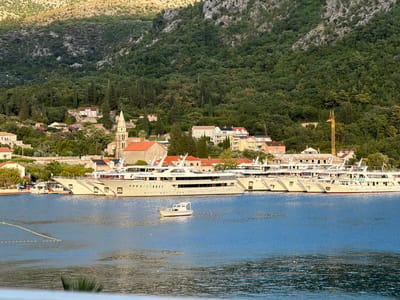 This image captures a scenic view of the coastline in Banići, Croatia, featuring a tranquil bay lined with moored yachts and a picturesque village nestled against a verdant, mountainous backdrop. The foreground is dominated by the shimmering blue waters of the bay, with a small white motorboat positioned centrally in the frame. Along the shoreline, a fleet of large, white yachts are docked, their sleek designs contrasting with the natural landscape.
Behind the yachts, the village of Banići unfolds. Buildings with red-tiled roofs are visible, interspersed with lush greenery and cypress trees. A prominent church steeple rises above the other structures, adding a historical and architectural element to the scene. The village is situated on a gentle slope, leading up to a densely forested mountain that forms the backdrop of the image. The mountain's slopes are covered in a thick canopy of trees, with patches of rocky terrain visible in the higher elevations. The sunlight casts a warm glow on the scene, highlighting the textures of the landscape and the details of the architecture. A construction crane is also visible among the buildings, suggesting ongoing development in the area. The overall impression is one of a peaceful, idyllic coastal community.