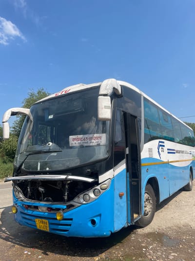 A blue and white Chandigarh Transport Undertaking (CTU) bus is shown in a partially damaged state. The front of the bus is visibly broken, with the hood open and internal engine components exposed. The license plate reads "CHO 1-G A1472". The bus route displayed on the front window indicates "Chandigarh 43-Dharamshala" and lists locations "Dehra, Vankhandi, Kangra, Gaggal". The background features a clear blue sky and some greenery, suggesting an outdoor location. The bus is parked on a roadside with a partially visible paved surface and some debris or dirt present.