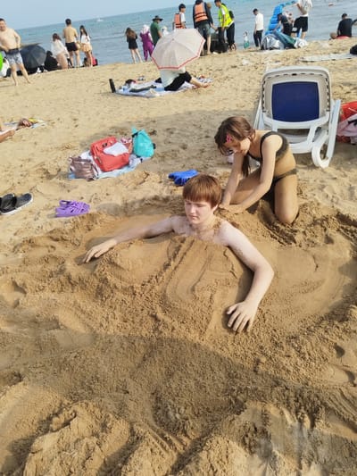 This media file captures a sunny day at a beach in Qinhuangdao, China. The foreground features two children partially buried in the sand. A young boy with reddish-brown hair is buried from the neck down, his arms resting at his sides. A young girl in a black bikini is kneeling behind him, her hands in the sand, appearing to be burying him further.
In the background, other beachgoers are visible, enjoying the day. Some are walking along the shore, while others are relaxing on the sand. One person is seated on a white beach chair. A collection of bags, including a red backpack and a blue tote bag, are placed on the sand near the children. Scattered on the sand are a pair of black Crocs and purple sandals. The ocean stretches out in the distance, with a few boats on the water. The lighting suggests it is daytime, likely late morning or early afternoon, with clear skies and warm weather conducive to beach activities. The overall mood appears relaxed and playful, with the children engaged in a common beach pastime.