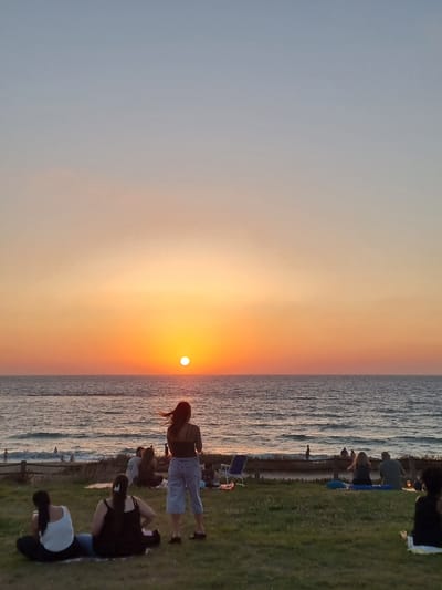 The image captures a sunset scene at the beach in Palmachim, Israel. The sun, a bright orange orb, is low on the horizon, casting a warm glow across the sky and the sea. The sky transitions from a pale blue at the top to shades of orange and pink closer to the sun. The sea is calm, with gentle waves rolling towards the shore.
In the foreground, several people are seated on the grassy area overlooking the beach. Two women in the lower left are sitting on the ground, one in a white tank top and the other in a black tank top. To their right, a woman in a black tank top and light blue shorts stands facing the sea. Further back, more people are scattered across the grass and on the beach, some sitting on blankets and chairs, watching the sunset. A few individuals are visible in the water.
The environment is natural, with a grassy slope leading down to a sandy beach bordered by the sea. The overall atmosphere is peaceful and serene, typical of a sunset gathering by the coast.