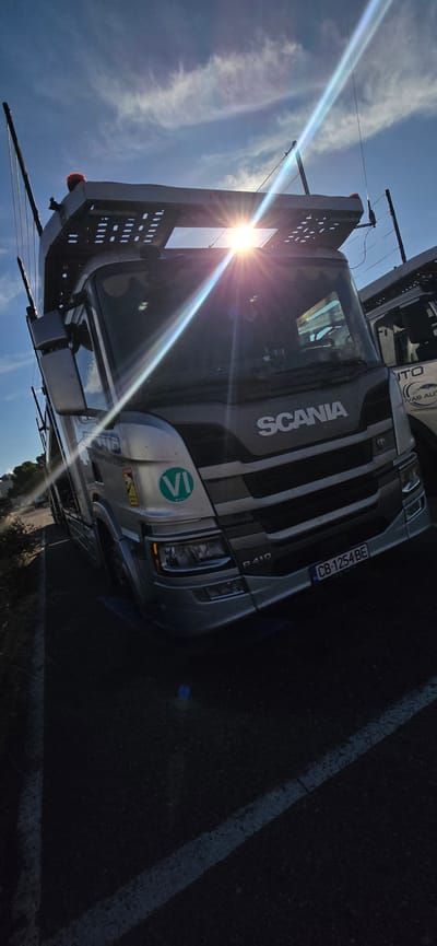 A silver Scania truck is parked on an asphalt surface, possibly a road or parking lot, with white lane markings visible. The truck is angled towards the right of the frame, with its front facing the viewer. The sun is bright and positioned in the upper center of the image, creating lens flare and overexposing the top of the truck's cabin. The sky is blue with some wispy clouds. The truck appears to be a car carrier, as evidenced by the structure on top designed to hold vehicles. A license plate on the front of the truck reads "CB 1254 BE". There is text on the side of the truck that is partially obscured by the sun and angle, but appears to include "Roto" and "SCANIA". Another vehicle is partially visible to the right of the main truck. The environment appears to be outdoors, with some vegetation visible on the left side of the frame and a building or structure in the background on the right. The presence of a "VI" sticker on the truck suggests it may be related to Italian vehicle regulations. The location is identified as Pedrola, Spain.