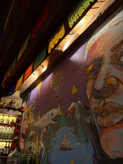 The image captures an interior view, likely a restaurant or bar, characterized by a vibrant and eclectic decoration scheme, primarily featuring a large, colorful mural on the ceiling. The photograph is taken from a low angle, looking upwards and diagonally across the room.
The most striking feature is the ceiling mural, which appears to cover a significant portion of the overhead space. In the upper right section of the image, a large, stylized face dominates, possibly that of a man or a mythical figure, with bold brushstrokes and earthy tones. The face has prominent eyes, a nose, and lips, and appears to be looking down.
Moving across the ceiling, the mural transitions to a purple background filled with various drawings and text. A central element here is the word "MEZCAL" written in green, accompanied by a stylized plant resembling an agave, and a skeletal figure below it. Other details on the purple background include small sketches, geometric yellow triangles, and an illustration of what appears to be an erupting volcano or mountain. Further along this section, a distinctive fish-like creature is depicted, standing upright or moving on two legs, with its mouth open and internal structures (possibly bones or muscles) visible. The overall style of the mural is artistic and somewhat surreal, blending figurative and abstract elements.
To the left and bottom of the image, a series of wooden beams runs diagonally, suggesting a structured ceiling or canopy. Strung between these beams are colorful paper cut-out decorations, characteristic of "papel picado," in hues of yellow, green, red, orange, and blue. Small, recessed lights are visible along the beams, casting light onto the decorations and the ceiling.
In the bottom right corner, a section of what appears to be a bar or shelf area is visible. It features several bottles, some empty and clear, alongside festive red decorative elements, possibly chili strings or garlands. Brass-colored bells or ornamental objects are also hanging in this area.
The lighting in the image suggests an indoor setting, possibly in the evening, with the small lights on the beams contributing to the ambient illumination. The tilted perspective of the photo adds dynamism to the scene, emphasizing the elaborate ceiling art and decorations.