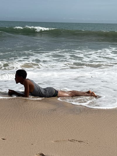A man is positioned head-down on a sandy beach along the coastline. His hands are placed on the sand, and his body extends upward into the white foam of the incoming waves. He is wearing a gray tank top and patterned shorts. The ocean stretches out to the left, with waves visible. Footprints are visible on the sand to the right of the man. The image shows Mopo-Akinlade, Nigeria.