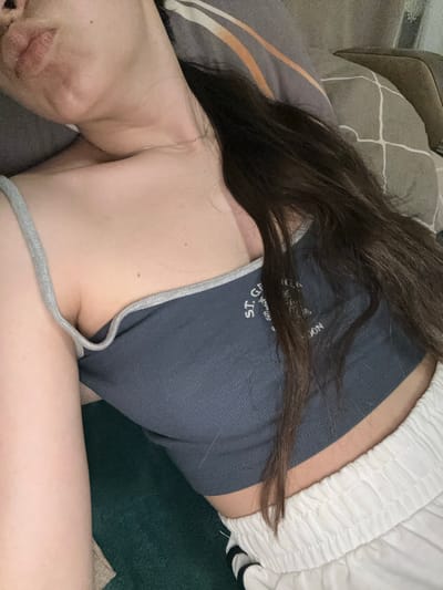 The image shows a close-up view of a person lying down. The person has long dark hair and is wearing a dark blue tank top with gray trim and white drawstring pants. The person is lying on a bed with a patterned comforter and pillows. The scene appears to be indoors. The provided text does not contain information about the city of Postoyata, Ukraine.