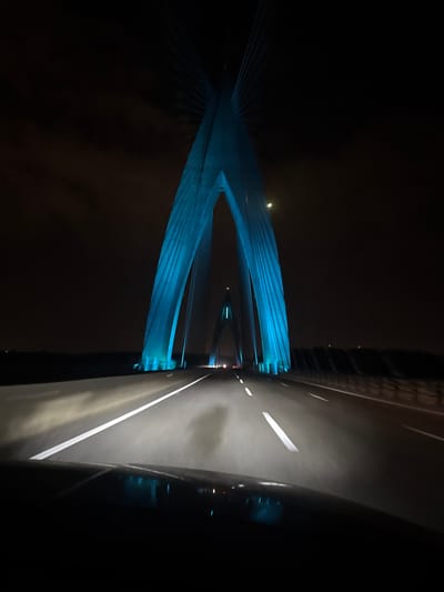 A nighttime view from a vehicle moving on a highway. A large, blue-lit bridge towers beside the road, its supporting cables fanning out into the dark sky. The road stretches into the distance, illuminated by vehicle headlights. Lanes are clearly marked. A car's side mirror is visible in the right foreground, reflecting the passing scene. The sky is dark with hints of mist or low clouds.