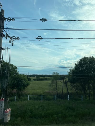 The image depicts a view through a window, likely from a moving vehicle, towards a rural landscape under a cloudy sky. In the foreground, a utility pole is visible on the left, with several stacks of cylindrical insulators hanging from it. Overhead, multiple parallel wires stretch across the frame, supported by what appear to be tensioners with pulley-like mechanisms. The wires are strung against a backdrop of a pale blue sky with wispy white clouds. Beyond the wires, a green field stretches into the distance, dotted with trees and some distant buildings. A white metal fence runs horizontally across the middle ground. The overall impression is one of a pastoral scene viewed from a mode of transport. The provided context places this scene in the city of Ushaki, Russia.
