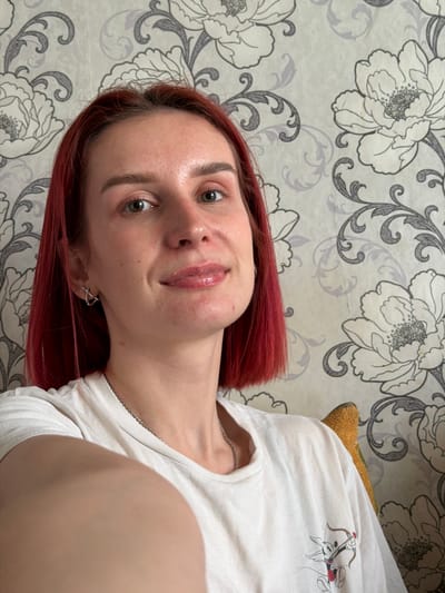 A young woman with red hair is taking a selfie indoors. She is wearing a white t-shirt and a silver necklace. She has fair skin, green eyes, and light eyebrows. The background is a wallpaper with a floral pattern in gray and white. The location is Заболотово, Russia.