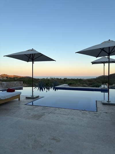 This image captures a tranquil outdoor scene in San José del Cabo, Mexico, during late afternoon or early evening. The foreground features a stone-tiled patio, upon which a light-colored wooden lounge chair with a white cushion rests on the left, holding a folded red towel. Two square, light-colored patio umbrellas with dark poles are positioned on the patio, one prominently on the left and another further back on the right, accompanied by a third, smaller umbrella behind it.
An infinity pool, lined with small blue tiles, dominates the midground, reflecting the sky. Beyond the pool, an arid landscape is visible, characterized by low-lying vegetation and distinct agave plants. The background reveals rolling hills with sparse greenery, leading to the horizon where the calm blue ocean meets a clear sky, transitioning from light blue overhead to warmer, yellowish tones near the horizon. A stone wall structure is discernible on a distant hill to the left. No human or animal subjects are present.