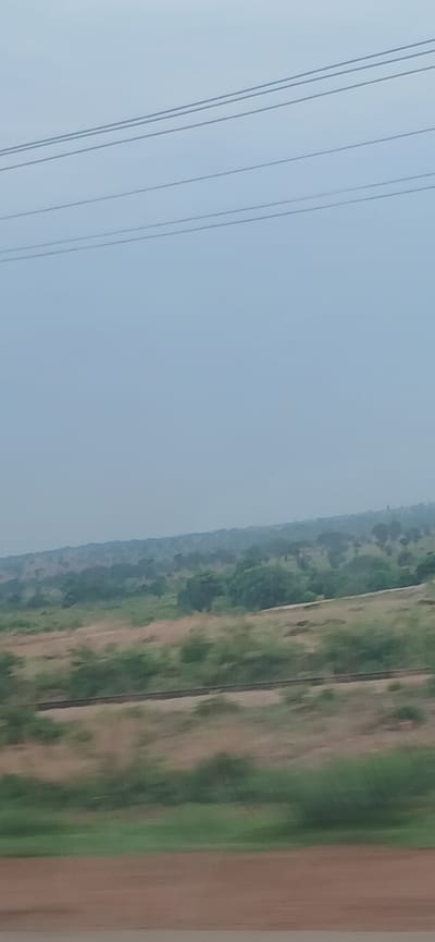 The image depicts a landscape view from a moving vehicle in what appears to be Kwarau, Nigeria. Power lines run diagonally across the top left portion of the frame. The background features a blend of green vegetation and trees, transitioning to a field or grassy area. At the bottom, a strip of brown or earth-toned ground is visible. The sky is a light blue. No people or other subjects are visible. The focus is on the scenery viewed from the vehicle's perspective.