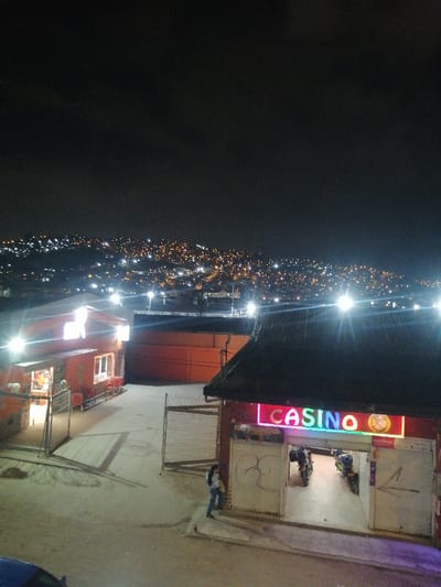 A nighttime shot reveals a sparsely populated area with buildings, including a casino with a brightly lit sign. A person stands near the entrance, and two motorcycles are visible inside. Streetlights illuminate the scene, casting shadows. In the distance, a hillside is dotted with countless lights, suggesting a sprawling urban area. The sky above is dark, hinting at a late hour.
