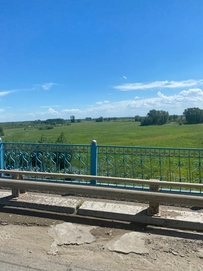 The image depicts a landscape scene viewed from a bridge or elevated walkway. The primary subjects are a blue wrought-iron fence and a concrete embankment. The fence, with its intricate scrollwork and decorative elements, runs horizontally across the middle ground of the image, separating the viewer from a vast green field. This field stretches into the distance, dotted with trees along its edges and forming the background. Above the field, a bright blue sky with scattered white clouds dominates the upper portion of the frame.
The scene appears to be an outdoor setting on a clear, sunny day. The shadows cast by the fence and embankment suggest it is mid-day. The location context of "Aqsu city administration, Kazakhstan" implies this is likely a public area, possibly a park or a roadside scenic overlook within the city. There is no activity or event taking place; the image captures a static view of the environment. Notable details include the weathered texture of the concrete and the stylized design of the fence. No text is visible in the image. The overall emotion conveyed is one of tranquility and a sense of open space.