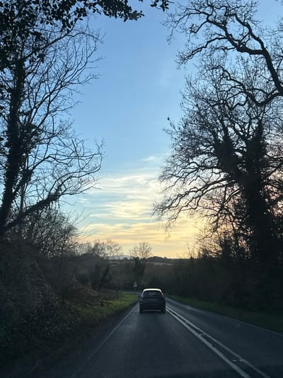 The image portrays a rural road scene within the Wyre Forest district, United Kingdom, during either dawn or dusk, characterized by a gradient sky from light blue overhead to yellowish-orange at the horizon. The perspective is captured from inside a vehicle, looking forward. A dark-colored hatchback vehicle, possibly black or dark grey, is centrally positioned on the asphalt road, traveling away from the viewer. The road features a continuous double white line down its center. Dense vegetation, consisting primarily of bare-branched deciduous trees and bushes, borders both sides of the roadway. Some trees on the left exhibit significant ivy growth. A small, white, triangular road sign with a red border is visible on the left verge in the mid-ground. Rolling hills and fields stretch into the distant background. No individuals are visible.