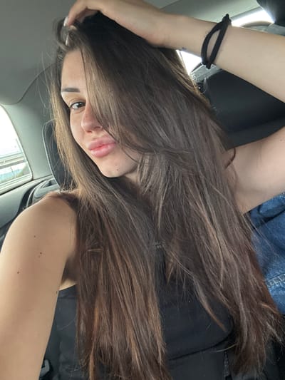 The image depicts a young woman inside a car, seen from a slightly high angle and rotated, giving a dynamic feel. Her long, voluminous dark brown hair, with subtle lighter brown highlights, dominates a large portion of the frame, flowing over her shoulders and chest.
She is looking towards the camera, with her left eye clearly visible, dark and engaging. Her nose and lips, slightly parted in a soft pout, are also in view. Her left arm is bent, with her hand near her head, and a black scrunchie is worn around her left wrist. A small, faint mark or tattoo is visible on her left forearm. She appears to be wearing a black top or garment, with a piece of blue denim fabric visible in the upper left background, possibly part of her attire or a jacket.
The setting is clearly the interior of a car, with elements like the headliner and blurred car window reflections visible. The lighting suggests it is daytime, with bright ambient light illuminating the scene. The overall impression is a casual portrait taken within a vehicle. There is no clearly discernible text within the image. The location context provided is Aldomirovtsi, Bulgaria.