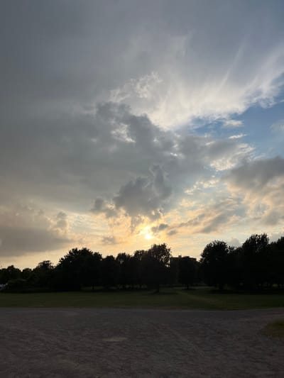 The image captures an outdoor landscape, likely a park or an open field, under a dynamic and varied sky. The scene is dominated by a expansive sky filled with numerous clouds. On the right side, the sun is visible, peeking through a break in the clouds and casting a bright, golden-yellow glow that illuminates parts of the sky and clouds with warm hues. This bright area contrasts with the left side of the sky, which is covered by darker, heavier grey clouds, suggesting mixed weather conditions or a transitional period.
Below the sky, a line of dense trees with dark, silhouetted foliage stretches across the mid-ground, appearing as a dark band against the brighter sky. To the left of the treeline is a strip of green grass, indicative of a field or lawn. To the right, a wider area of unpaved ground, possibly a dirt path or gravel road, runs parallel to the trees.
The time of day appears to be late afternoon or early evening, consistent with a sunset or sunrise, given the low position of the sun and the interplay of light and shadow on the clouds and landscape elements. No specific activities or events are taking place, and no people or visible text are present in the image. The overall impression is one of natural beauty with dramatic cloud formations and atmospheric lighting.