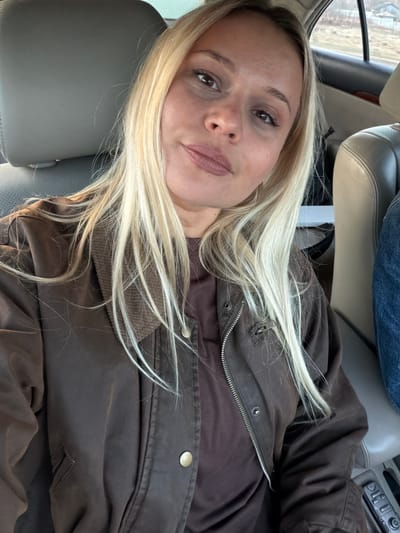 A blonde woman is sitting in a car. She is wearing a brown jacket and a brown shirt. The car seat is gray leather. The location is unknown and there is no evidence in the image to suggest that it is Дулесово, Russia.