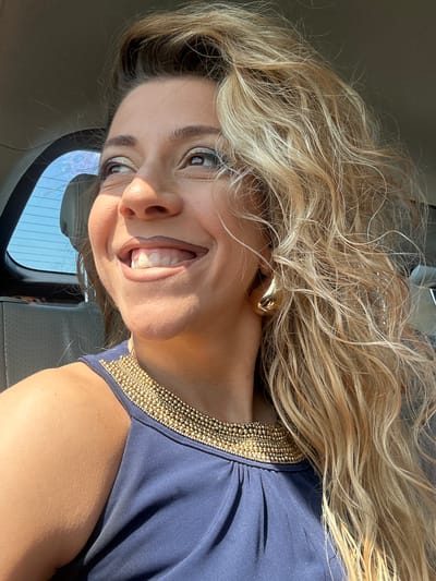 A smiling woman with long, blonde, curly hair is shown in a close-up, slightly angled portrait. She is wearing a blue dress with a gold beaded neckline and gold earrings. The woman is inside a vehicle, based on the visible car window and interior. The location of Lelystad, Netherlands, is not discernible from the image.
