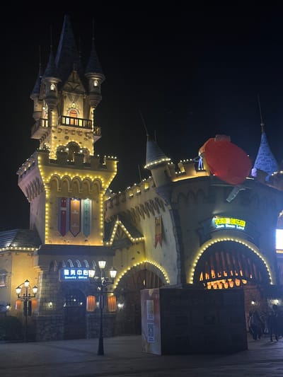 An illuminated castle-like building dominates the night scene. Its architectural details include conical turrets and intricate window designs. The building is adorned with strings of lights, highlighting its structure. A large red balloon-shaped object is positioned near the upper portion of the building. The image captures the magical atmosphere of the location.