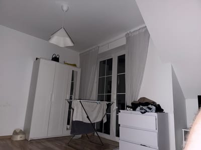 In Falenty Nowe, Poland, a bedroom scene is depicted. A white wardrobe stands to the left, with a small beige potty at its base. In the center, a clothes drying rack is set up, holding damp laundry, including a grey towel and dark fabric. Behind the drying rack are double glass doors, draped with light grey and white striped curtains. To the right of the doors, a white chest of drawers is visible, with a pile of folded clothes on top. The room features white walls and a sloped ceiling, with a white pendant light fixture hanging from above. The floor is covered in light brown wooden planks.