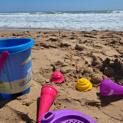 The image shows a collection of colorful plastic beach toys scattered on sandy ground, likely on a beach. The toys include a blue bucket, a pink funnel, a purple sifter, a pink flower-shaped mold, a yellow spiral-shaped mold, and a purple cone. Some of the toys are partially buried in the sand. Behind the toys, the beach extends to the ocean, where white waves crash onto the shore. The image is taken in Guardamar Del Segura, Spain.