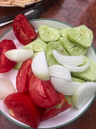 A close-up, overhead shot shows a white plate with a green rim, filled with sliced tomatoes, cucumbers, and white onions. The vegetables are artfully arranged on the plate. In the upper left background, a portion of another dish, possibly pasta, is visible, along with a fork and spoon. The plate rests on a wooden surface. The lighting is natural, casting soft shadows. There are no people visible in the image. The setting appears to be a mealtime scene. No specific location in Muhametjan Tüymebaev, Kazakhstan, is identifiable from the image.