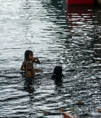 Two children are playing in murky water near a red structure, possibly a boat. The child on the left is sitting in the water, holding a piece of wood or debris, and appears to be eating something. The child on the right is swimming. The water is reflective and shows ripples. Debris is visible floating on the surface and along the edge of the water in the foreground. The location is identified as Soutr Nikom, Cambodia.