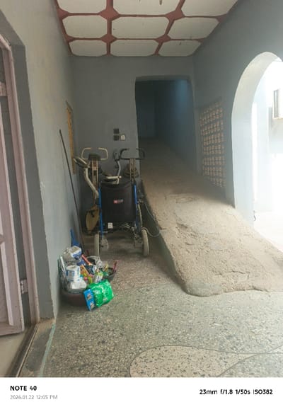 This image displays a semi-covered passageway located in Gwammaja, Nigeria, captured on January 22, 2026, at 12:05 PM. The area features light grey-painted walls and a white ceiling adorned with a reddish-brown octagonal grid pattern. The floor is terrazzo-style, exhibiting light grey and beige tones with a distinct, curved demarcation line.

On the left, a light-colored door frame is partially visible. A tall, dark pole leans vertically against the wall, below a high-set, narrow window with a yellow tint. At floor level, a black basin contains miscellaneous household items including toilet paper rolls, plastic containers, and small packages; a green plastic bag rests nearby.

Adjacent to the left wall and preceding a substantial concrete ramp, two pieces of equipment are present: a stationary exercise bike with white handles and a beige frame, and a blue-framed rollator or wheeled walker featuring a black fabric seat/storage compartment. Above these, two small, dark electrical boxes are affixed to the wall.

The central feature is a wide, upward-sloping concrete ramp, textured with loose sandy material, that leads into a darker, unseen area. To the immediate left of the ramp, a dark arched doorway opens into a dim interior. To the right of the ramp, a larger archway provides access to a brighter exterior space, where a rectangular vent is visible on a distant white wall and a reddish-brown patterned grate is integrated into the wall adjacent to the ramp. No individuals are visible within the frame.