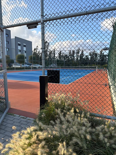 The image depicts an outdoor tennis court in Tserkezoi, Cyprus, viewed through a chain-link fence. The court has a red-orange surface with a blue area in the center. To the right of the court, tall grass and plants grow along a brick walkway. A modern, multi-story building with grey walls is visible in the background beyond the fence. The sky is blue with scattered clouds. A black structure partially obstructs the view of the court's net. The image appears to be taken from a low angle, looking up at the tennis court.