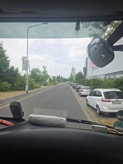 The image, taken from inside a vehicle, shows a view of a street lined with parked cars and trees. The time of day appears to be daytime, under a cloudy sky. The scene is set in Schweinfurt, Germany, as indicated by a license plate on a white van reading "SW LC 304".

Prominently featured are several white and silver vans parked along the side of the road. A large building with some signage is visible in the background, with a billboard that is partially obscured. The interior of the vehicle shows a portion of the dashboard, including a roll of paper towels, a dashcam, and a side-view mirror reflecting the sky and trees. A green sign with text is also visible along the roadside. The overall impression is a snapshot of a typical urban or suburban street on a possibly overcast day.