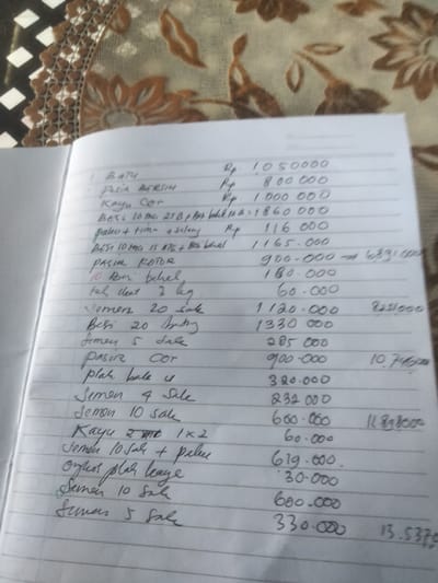 The image shows a handwritten list in a notebook. The notebook is open and lying on a surface with a patterned covering. The list appears to be an itemized bill or record of purchases, with quantities and costs next to each item. The costs are marked with "Rp," which typically denotes Indonesian Rupiah. Some items on the list include building materials such as "Batu" (stone), "Pasir" (sand), "Kayu" (wood), "Besi" (iron), "Semen" (cement), and other related items. Some quantities and descriptions include "20 sak" (20 sacks), "5 sale," and size measurements like "1x2" for lumber. Some totals or calculations are written to the right of the itemized list. The city is not mentioned in this image.