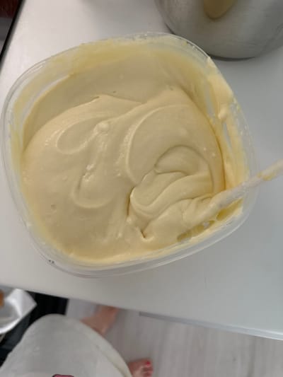 An overhead shot captures a clear plastic container filled with a thick, pale yellow batter. A white spatula with some batter clinging to it is inserted into the mixture, creating swirls and peaks. The batter appears smooth with some small bubbles visible. To the left of the container is a portion of a silver bowl. The background is a white surface, possibly a countertop, with a sliver of a person's foot with pink nail polish visible on the right side of the frame, suggesting a home setting. The lighting appears bright and even, indicating it is daytime. The image does not contain any visible text.