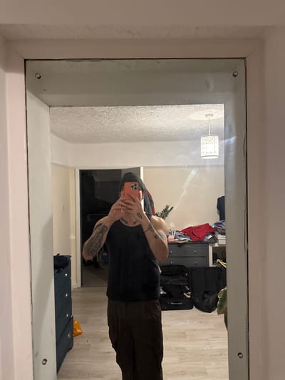 A man stands in front of a large, rectangular mirror, taking a photo of himself with a phone. He wears a dark t-shirt and brown pants, a beanie, and his arms are covered in tattoos. The room appears cluttered, with suitcases, clothes, and a dresser visible in the background. The location is possibly a residential space in Hitchin, United Kingdom, based on the style of the interior and typical UK household items. The mirror is attached to the wall with visible screws.