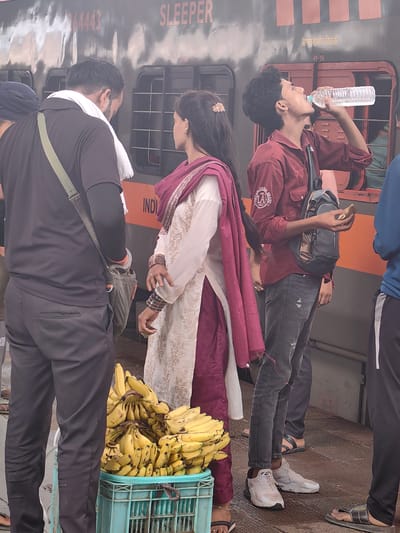 At the Itarsi railway station, a group of individuals is captured in a candid moment near a train. A young man, dressed in a maroon shirt and distressed denim jeans, is drinking from a clear plastic water bottle. He wears a grey sling bag across his chest. Beside him, a woman in traditional Indian attire, a white and maroon salwar kameez, stands with bangles adorning her wrist. To their left, a man in a black t-shirt and black pants, with a white scarf draped around his neck, is seen from the side. In the foreground, a crate overflowing with ripe yellow bananas is visible. The train, with "SLEEPER" written on its side, forms the backdrop. The overall scene depicts a snapshot of daily life at the bustling railway station.