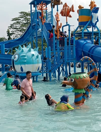 The image captures a vibrant scene at a water park in Durin Simbelang, Indonesia. The setting features water playground structures adorned with cartoonish sea creatures, including an orange coral, a seahorse, a striped fish, a dolphin, and a pufferfish. A multi-tiered blue water slide structure rises in the background. Several people are enjoying the water, including adults and children. In the foreground, a man wearing a pink shirt is seen wading in the water with two young girls. Nearby, another girl is swimming toward an interactive water feature shaped like an elephant. A woman wearing a hijab sits in a yellow inflatable ring. Others can be seen spread throughout the pool, some swimming and others standing. The water appears shallow and the overall atmosphere is one of lively recreation.