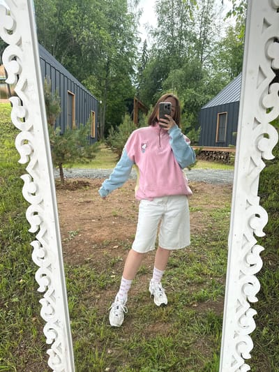 The media file shows a person taking a mirror selfie outdoors. The person, who appears to be a young woman with brown hair, is wearing a pink and light blue color-blocked quarter-zip sweatshirt with a Hello Kitty graphic, white shorts, white athletic socks with a pink pattern, and white sneakers. She is holding a smartphone in her right hand.

The setting appears to be a backyard or rural area. In the reflection of the mirror, two dark-colored, modern-looking cabins or houses with metal roofs and large windows are visible. The surrounding environment consists of green grass, dirt, gravel, and lush trees. The sky is overcast, suggesting a cloudy day. The mirror itself has an ornate white wooden frame with carved decorative elements.

The overall scene conveys a casual, relaxed mood. There is no visible text in the image. Based on the architecture and natural surroundings, the location is consistent with Novaya Vozhoika, Russia.