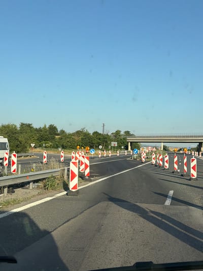 The image captures a clear, sunny day scene on a highway, likely within or near Kaspichan, Bulgaria, characterized by road construction. The foreground shows a paved road with a white lane marker. Numerous traffic cones and temporary road barriers, primarily red and white striped pylons, are strategically placed to redirect traffic. Several traffic signs are visible, including directional arrows and regulatory signs indicating road work ahead. A metal guardrail runs along the left side of the road, separating it from a grassy shoulder. In the background, a concrete overpass spans the highway, and a power line tower is visible in the distance. The environment is bordered by lush green trees and vegetation on the left and right sides of the road, suggesting a rural or semi-rural setting. The sky is a vibrant, cloudless blue. No people are directly visible in the frame, but the presence of the construction equipment implies human activity related to road maintenance or repair. The overall impression is one of ongoing infrastructure work on a major roadway.