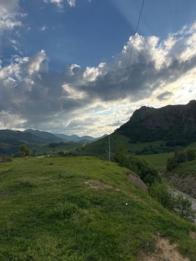 This is a vertical, eye-level, medium shot of a mountainous landscape under a partly cloudy sky. The foreground is dominated by a lush, green grassy slope. To the right of the frame, the slope drops off into a narrow, unpaved road or track running alongside a steep, eroded embankment covered in vegetation. Further back, rolling green hills covered in trees recede into the distance, culminating in a series of hazy blue mountain ranges. The sky is a vibrant blue, with dramatic clouds, some of which are backlit by the sun, creating rays of light. A single thin power line stretches diagonally across the upper right portion of the frame. There are no people or animals visible in the image. The scene appears to be in a rural, undeveloped area. The geographical location is the city of Nizhnyaya Mara, Russia.