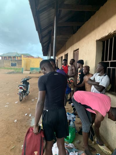 The image depicts a group of men standing outside a building with a covered walkway in Kagwaru, Nigeria. The building appears to be constructed with stucco walls and a wooden roof structure. Several men, most of whom are shirtless or wearing casual attire, are gathered near the wall. Some are looking at each other, while others appear to be occupied with personal items. In the foreground, a man in a black t-shirt and shorts is holding a maroon backpack. To the left of the building, a white and black motorcycle is parked on the dirt ground. Scattered debris and a green plastic bag are also visible on the ground. In the background, there are structures that suggest a more developed area, with a yellow and green building and another building with a sloped roof. The sky is overcast, indicating potentially cloudy weather.