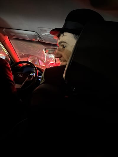 A person with light skin and a dark beard is wearing a black hat inside a car. The person is looking toward the left side of the frame. The car's interior is visible, including the steering wheel with the Toyota logo, dashboard, and window. Red and white lights are visible outside the car window. The image appears to have been taken at night. The location in the city of Fort Lee, United States of America, is not discernible from the image.
