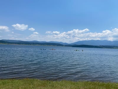 This media file depicts a scenic view of Batak Reservoir in Bulgaria under a bright, partly cloudy sky. The foreground is dominated by a grassy bank sloping down to the water's edge. The reservoir itself occupies the majority of the frame, its surface rippled by a gentle breeze. In the mid-ground of the water, three separate groups of people are engaged in kayaking. The first group, on the left, consists of two kayakers in a single blue kayak. To their right, another kayak, orange in color, carries a single person. Further to the right, a third kayak, green, has one individual paddling. The background features rolling hills and mountains, covered in lush vegetation, receding into the distance. The time of day appears to be daytime, likely late morning or early afternoon, given the bright sunlight and clear blue sky with scattered white clouds. The overall mood is peaceful and recreational, suggesting a pleasant day spent outdoors enjoying water activities. There is no visible text in the image.