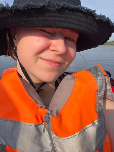 The image is a close-up selfie of a young person wearing a black bucket hat and an orange life vest. The person is looking directly at the camera with a slight smile and one eye closed, suggesting a wink or squint. The background shows a body of water with a faint shoreline in the distance. The lighting indicates it is daytime, with the sun casting a warm glow on the person's face. The scene appears to be on a boat or near the water, with the life vest suggesting recreational activity. The location is described as being in Kalino, Russia, implying an outdoor setting by a river, lake, or sea.