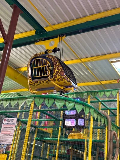 The image displays an interior view of an indoor amusement park or play area, identified as Actiland, located at Espace commercial des Prairies, 14970 Bénouville, France. A yellow ride vehicle, designed with brown giraffe-like spots and resembling a compact helicopter cabin, is suspended from an overhead yellow rail system. A single individual is discernible inside the cabin, partially obscured by yellow horizontal bars. Below the suspended vehicle, a multi-level soft play structure is visible, constructed with yellow support poles and green safety netting. The top edge of this structure is decorated with a green border featuring numerous light-green, leaf-shaped fabric elements. On the lower left, a white sign with red and black text in French clearly reads "REGLEMENT AIRE DE JEUX ACTILAND/PLAYLAND". The ceiling, composed of corrugated metal panels, is supported by a framework of yellow and dark green metal beams, with a brown vertical support column positioned on the left side of the frame.