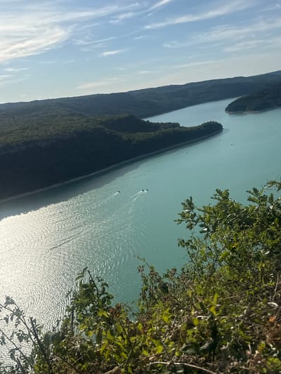 A high-angle, sunlit shot shows a wide, turquoise lake bordered by dark green, tree-covered hills under a pale blue sky with wispy clouds. In the foreground, dense green foliage and branches frame the lower right corner. Three motorboats create white wakes on the water, moving away from the viewer toward the center of the lake. A sailboat is visible further in the distance on the right side of the lake. The lake appears to be the Lac de Vouglans, located in the Jura department of France, near the commune of Moirans-en-Montagne.