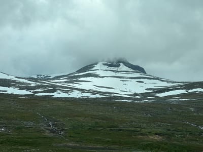 This image depicts a mountain landscape under a cloudy sky. A large mountain dominates the background, with its peak obscured by mist and clouds. The mountain is partially covered in snow, with patches of dark rock visible. In the foreground, there is a gently sloping terrain covered in green grass and scattered rocks. A small stream or watercourse winds its way down from the snow-covered upper slopes through the grassy area. The overall impression is one of a remote and rugged natural environment. The location is identified as Storforshei, Norway.