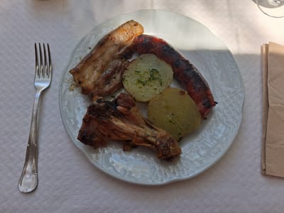 A close-up, overhead shot shows a white plate filled with grilled meat and potatoes, set on a white tablecloth with a diamond-shaped pattern. A silver fork rests to the left of the plate. The meal consists of a piece of grilled pork belly, a reddish-brown sausage, a chicken leg, and three round slices of potato, seasoned with chopped green herbs. The food appears to be a hearty, rustic dish. Sunlight streams in from the upper right, casting shadows across the plate and the tablecloth, suggesting it might be daytime. The setting appears to be a restaurant or a home dining area. No text is visible in the image. The overall impression is of a simple, satisfying meal.