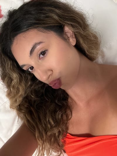 A young woman with dark, curly hair and brown eyes lies on a white surface, possibly a bed, with her head tilted towards the camera. She is wearing an orange top and has her lips puckered in a kissing gesture. Her skin appears tanned and dewy. She has a small hoop earring in her left ear and a stud earring. The background is a blurred white surface. The image appears to be a selfie taken in a bedroom. There is no indication of the city of San Juan, United States of America, in the image.
