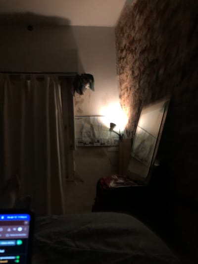 A dimly lit indoor scene, potentially in Beccar, Argentina, shows what appears to be a basement or storage area. The ceiling is low and partially covered with rough, possibly insulated material. A window or skylight is visible near the ceiling, providing a source of light augmented by a small lamp. Stairs lead up to a higher level. An object, possibly a hat, sits atop a structure near the wall. A person is present in the frame, partially obscured on the right. In the foreground, a mobile phone screen displays audio editing software.